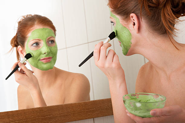 Young woman using mirror to apply a green facial mask stock photo