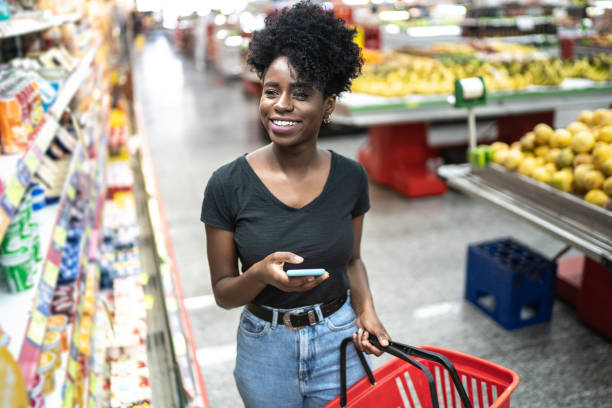young woman using mobile phone and choosing product in supermarket - natural food web imagens e fotografias de stock