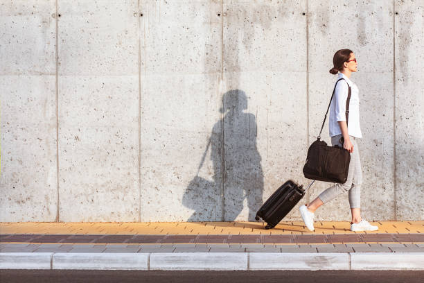 Young woman walking on a sidewalk beside the concrete wall and pulling a small wheeled luggage stock photo