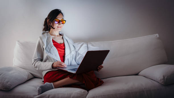 Young woman wearing blue light blocking glasses (yellow lenses) working with laptop on white sofa, health and wellness concept for melatonin and circadian rhythm stock photo