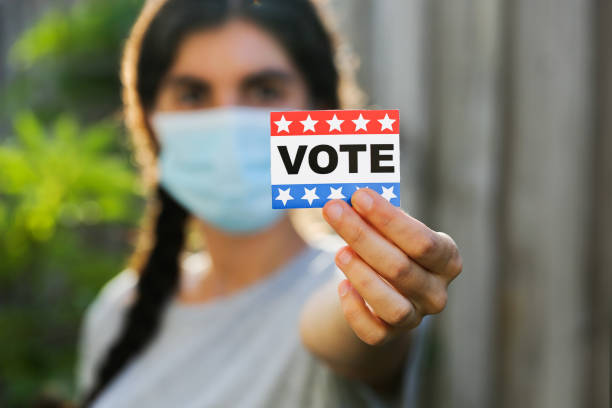 Young woman wearing face mask holding Vote sticker.