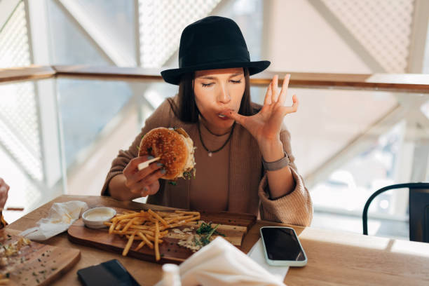 A young woman with a hat on her head sits alone at a table in a restaurant and enjoys unhealthy food. stock photo