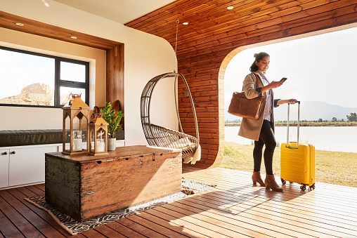 Young woman with a suitcase using her mobile phone while standing at lounge of her vacation home during a trip