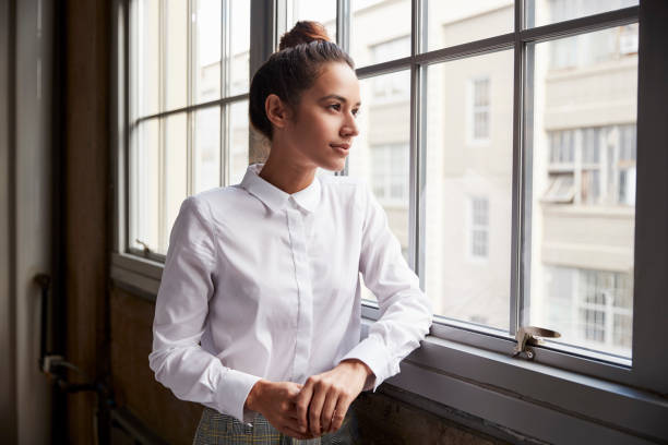 Young woman with hair bun looking out of window, waist up Young woman with hair bun looking out of window, waist up blouse stock pictures, royalty-free photos & images