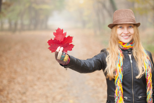 Young blond woman with hat, multicolored scarf and fingerless gloves holding red leaf. Looking at camera.