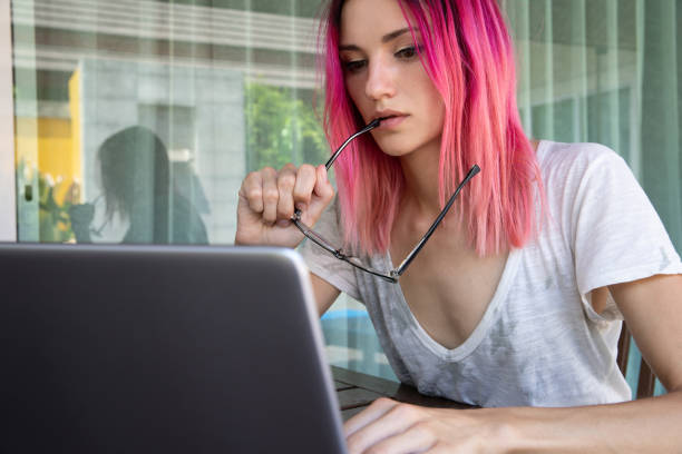 Young woman with pink hair working with laptop in cafe or home terrace. Young woman with pink hair working with laptop in cafe or home terrace. Serious pensive hipster girl freelancer work hard. pink hair stock pictures, royalty-free photos & images