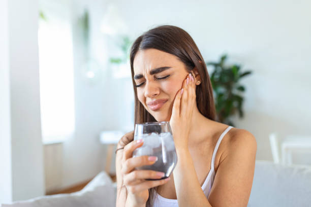 Young woman with sensitive teeth and hand holding glass of cold water with ice. Healthcare concept. woman drinking cold drink, glass full of ice cubes and feels toothache, pain stock photo