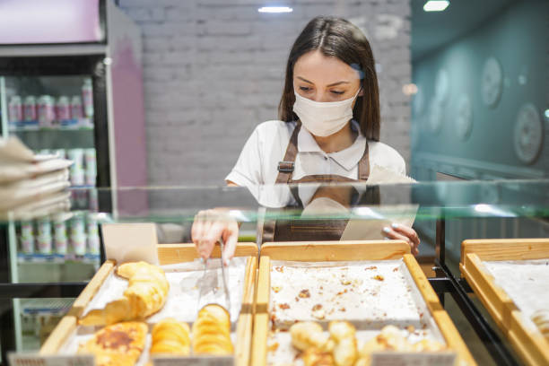 Young woman working at bakery and picking fresh pastry from display cabinet Young woman working at bakery and picking fresh pastry from display cabinet display cabinet stock pictures, royalty-free photos & images