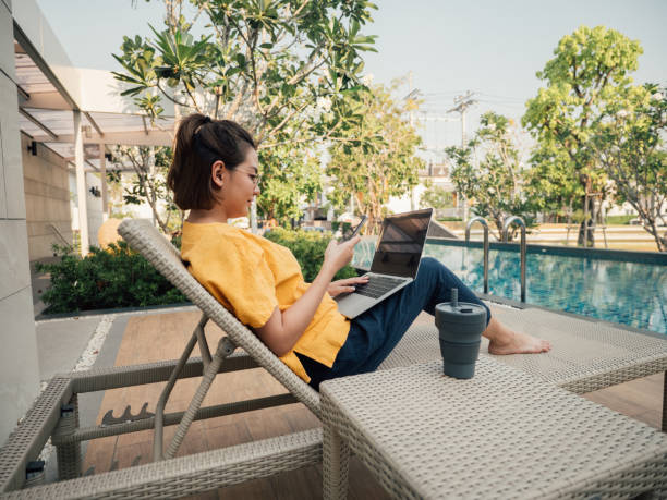 Young woman working on her computer on vacation near pool. Young asian businesswoman using smart phone and typing on laptop computer for sharing social media sitting in bed at outdoor nomadic people stock pictures, royalty-free photos & images