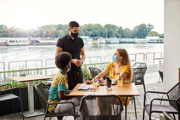 Young Women Enjoying In Restaurant - New Normal Friendship Concept Two young women sitting in restaurant and talking with waiter. All wear protective face masks. waiter taking order stock pictures, royalty-free photos & images