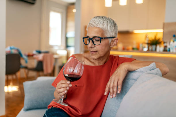 You've gotta spoil yourself every now and then Photo of mature sad woman sitting on sofa with one glass of red wine waiting for somebody (man) - loneliness, quarrel, celebration Valentine Day alone concept. Lonely mature woman holding glass of alcoholic drink while sitting on sofa at home during the day. alcohol abuse stock pictures, royalty-free photos & images