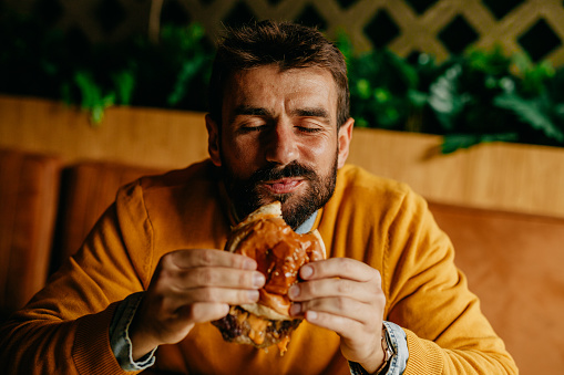 Spontaneous and cute portrait of a handsome enjoyable looking bearded man, having a bite of a cheeseburger and facing the camera with a big yummy smile and eyes closed. Looking and feeling like a kid when I eat this