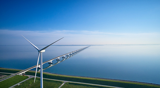 The Zeelandbrug aerial with windturbine during sunrise. The Sky is blue and it’s reflected in the water of the North sea. The bridge is also reflected in the water. Cars drive at a high speed over it.