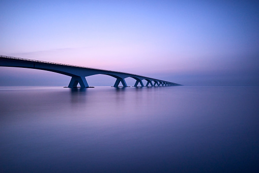 The Zeelandbrug on a cloudy day. The sky is purple and blue and is reflected in the water.