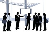 A vector silhouette illustration of business associates watching a monitor on which a mature man in a suit is giving a presentation.