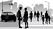 A vector silhouette illustration of people in a city.  People walk across a side walk while a car waits to turn.