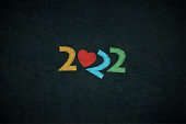 Creative fun horizontal vector wallpaper or poster or banner with text year 2022 in pastel colourful stroking pattern over scratched grunge textured black backdrop like embroidery or colorful writing on a chalkboard. There is no people and copy space. Apt for use as happy new year banners, posters, greeting cards and gift wrapping paper sheets. The heart and and a2 are tilted in a cute funny way.
