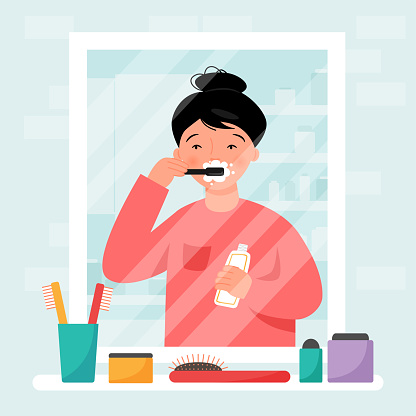 Girl brushing her teeth in the bathroom. The girl is reflected in the mirror. Bathroom interior. Shelf with toiletries. Routine hygiene.