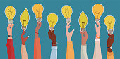istock Raised arms of diverse and multi-ethnic business people holding a light bulb shaped label as a concept of innovation or startup or collaboration or financial investment.Community concept 1339256703