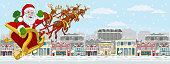 Christmas street scene with Santa Claus in his Christmas sleigh with reindeer flying over Victorian style houses, shops and other buildings in the snow.