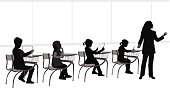 Silhouette of a teacher standing and talking in front of a second grade classroom with four students sitting at their desks.  Some of the kids are writing, some reading and some listening to their teacher.