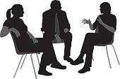 A vector silhouette illustration of a group of three people engaged in discussion incliding a young man speaking and gesturing, and a mature man and young woman listening.  They sit on chairs.