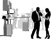 A vector silhouette illustration of people working near a stand-up desk using their smartphones. A business man talks onthe phone and a young woman texts near a desk where there is a printer, computer, monitor, filing cabinets, and bookshelf.