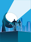 A vector silhouette style illustration of a couple of superheroes on a building rooftop with big searchlight and city skyline in the background. Put your logo or text on the searchlight or in other space available.