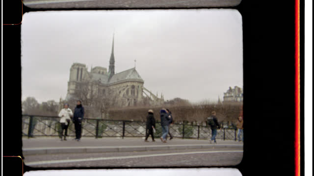 16mm. Bolex film footage. View of the Notre Dame Cathedral in Paris, France as tourists and locals cross a cobblestone bridge over St. Joseph River.