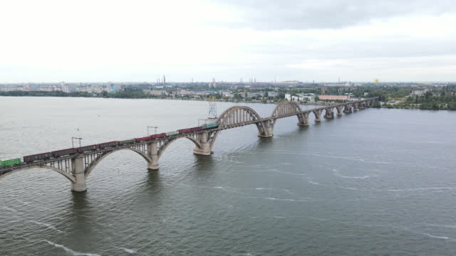 Aerial view of a train crossing a beautiful bridge. Freight train moves along the railway bridge. Aerial view of the old arched railway Merefo-Kherson bridge in the city of Dnipro.