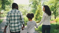 istock Back view of smiling dark-skinned man and woman walk in the park holding the hands of their little daughter. Being together, family weekend, picnic in the park 1323157431