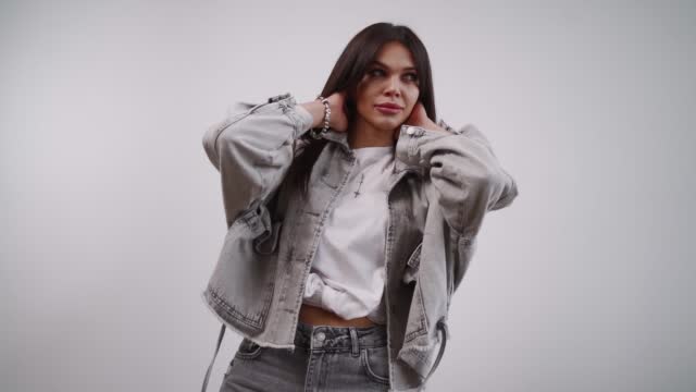 Beautiful stylish brunette woman in a denim jacket posing while developing her hair in front of the camera on a white background. The concept of modern fashion and style. Slow motion