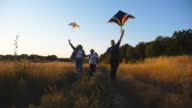 istock Close up of mommy and daddy with little daughter jogging with rainbow kites on rural road at sunset. Young parents with small cute girl holds hands of each other running among trail near grass field. 1370915212