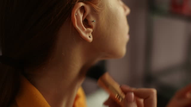 Close-up cropped shot of cute redhead young woman looking in mirror, putting powder on neck with big professional makeup brush.