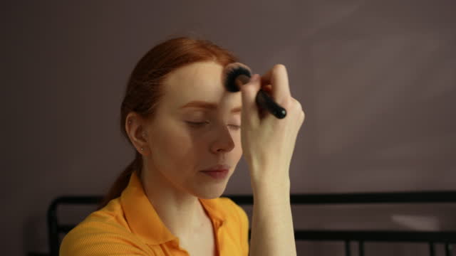 Close-up side view of serious redhead young woman applying cosmetic powder with big professional makeup brush, looking in mirror.