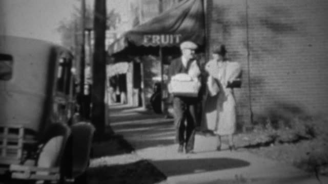 1936: Couple grocery shopping from fruit store walking bags of food.
