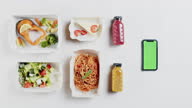 istock Food delivery app top view, take away meals in disposable containers on white background. Close-up of lunch boxes with cooked salad, spaghetti and cakes, portions. Smartphone with chroma green screen. 1369981173