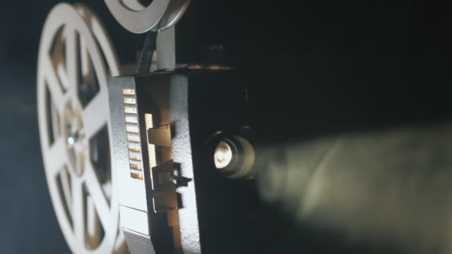 Front view of an old-fashioned antique Super 8mm film projector, projecting a beam of light in a dark room next to a stack of unraveled film reels