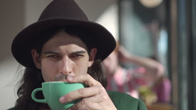 Headshot portrait of stylish retro man with long hair in hat smelling and drinking freshly brewed coffee from cup. Close-up of satisfied young Caucasian dandy resting in coffee shop indoors in 1980s.