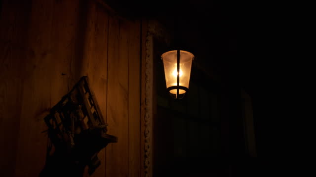 Iron lantern swaying in wind, night light, shadows on wooden wall of rustic barn. lonely lantern on dark street. Yellow light illuminates wall of building, wind sways it, shadows run along wooden wall, ominous atmosphere, warm light of lantern
