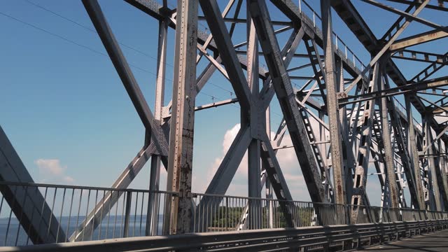 metal bridge. rough iron constructions against blue sky and river, filmed in motion from a car. side view. Railway and road bridge across the river. Silhouette of crossing steel beams
