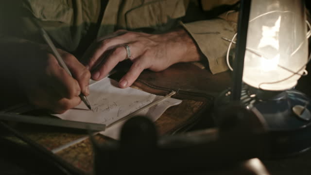 Soldier writing letter to home. Tilt down view of Soviet soldier sitting at table and writing message to home in dim light of oil lantern while taking break during World War II