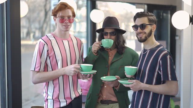 Three positive young Caucasian retro men in sunglasses clinking coffee cups drinking and smiling looking at camera. Portrait of happy relaxed friends resting in coffee bar in 1980s 1990s posing.