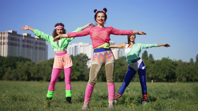 Wide shot slim fit retro woman with hair buns exercising with friends outdoors on sunny meadow. Happy Caucasian fitness instructor training young ladies outdoors in 1990s in summer park.