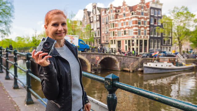 Woman vacationing in Amsterdam, taking pictures of canals with retro camera, smiling into camera