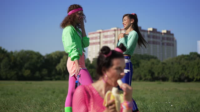 Women discussing blurred friend eating banana laughing mocking in slow motion. Portrait of retro Caucasian friends gossiping making fun of woman outdoors on sunny day. Lifestyle and 1990s concept.
