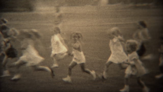 1937: Young girls foot race across grass park field in formal white dress.