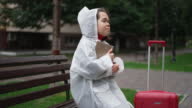 istock Young little person looking up hugging book sitting on bench with suitcase. Portrait of Caucasian little woman in rain coat enjoying hobby travelling in city outdoors. Slow motion. 1404230502